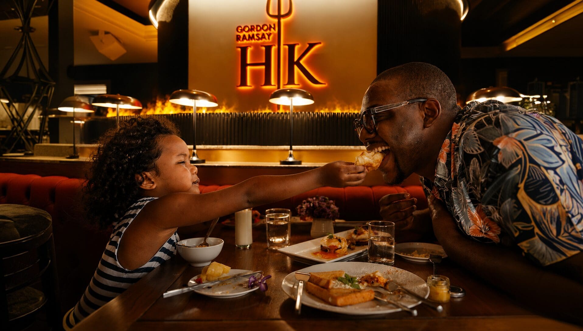 A child feeds a man at a restaurant with a “Gordon Ramsay HK” sign glowing in the background.
