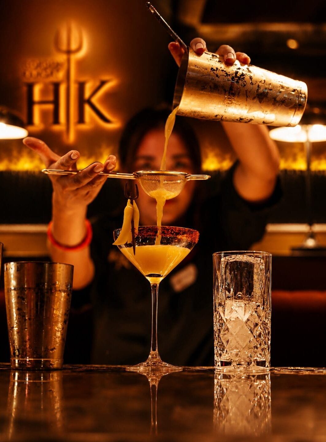 A barman strains a yellow cocktail into a glass at a dimly lit bar with an HK logo in the background.