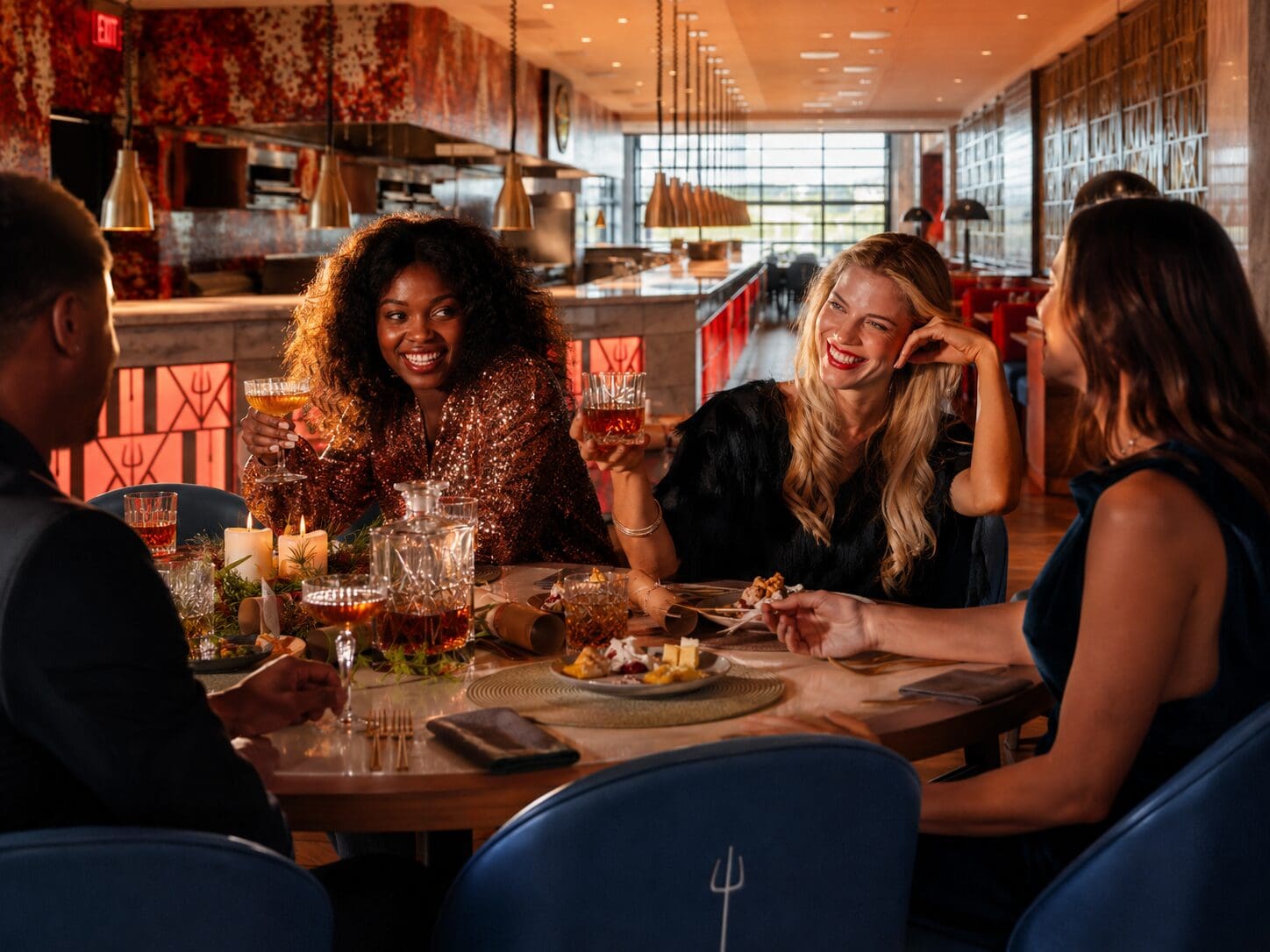 Four people dressed up, smiling and clinking glasses at a stylish restaurant table with food.
