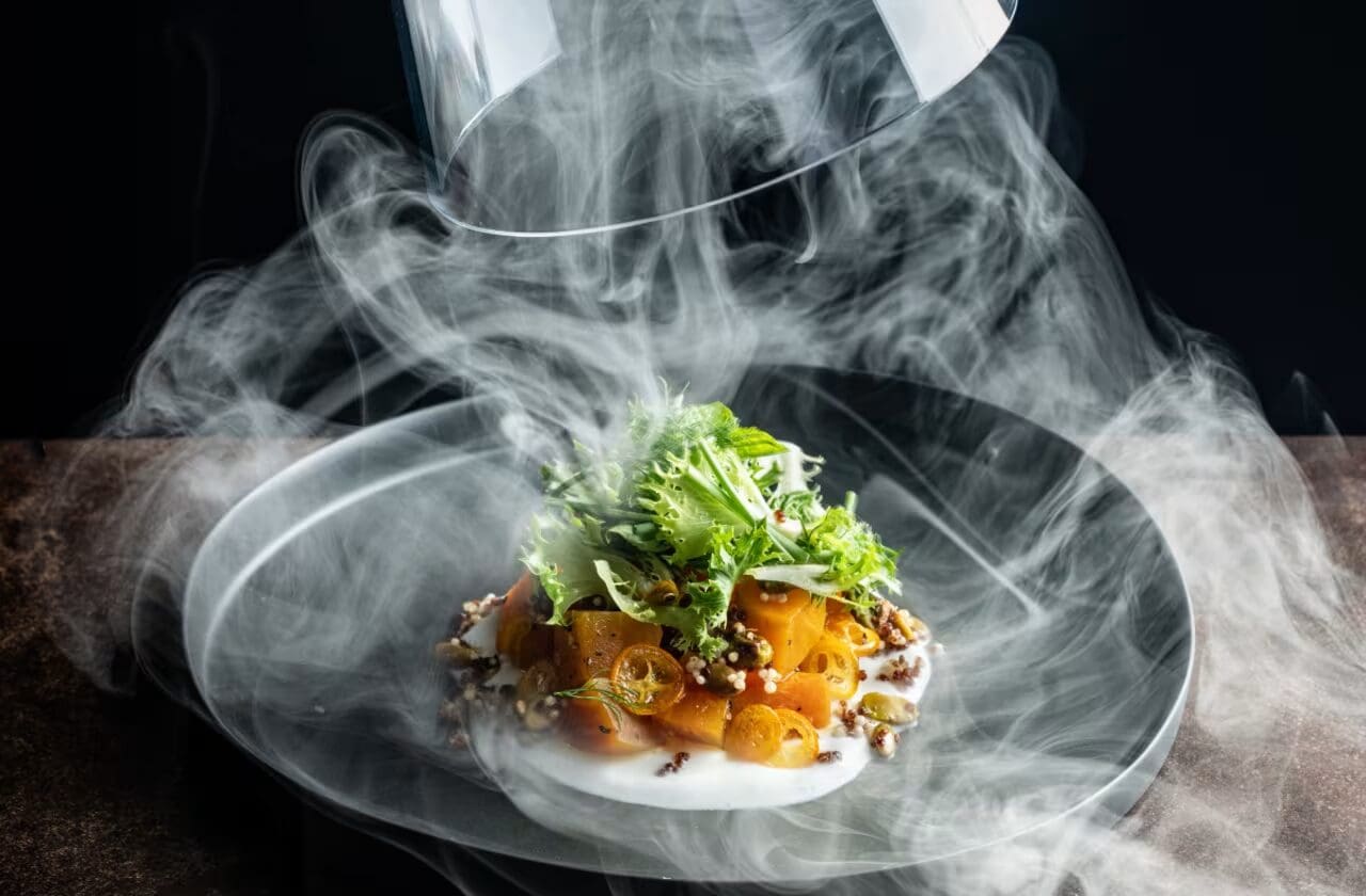 A gourmet salad with leaves and orange tomatoes under a glass dome with swirling smoke.