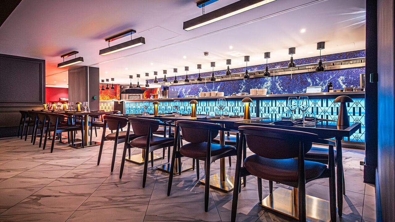 Modern restaurant with dark chairs, tables, and a glowing blue bar, with warm and stylish lighting overhead.
