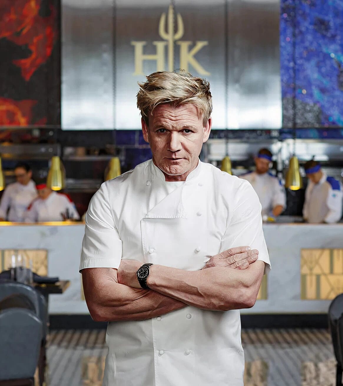 Gordon Ramsay in a white uniform stands with arms folded in a modern restaurant kitchen with staff in the background.