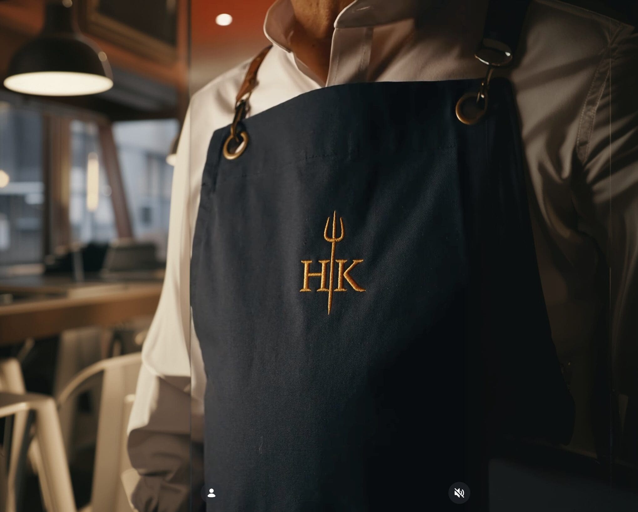 Person wearing a dark apron with “HK” and a trident logo, standing in a modern restaurant setting.