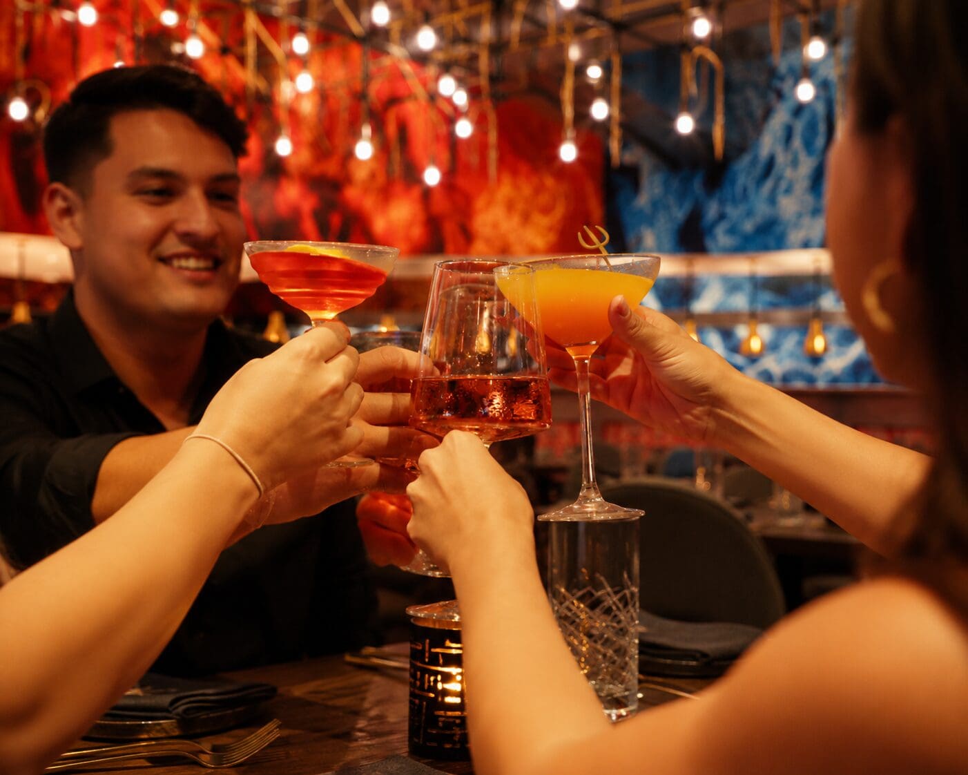 Four people clink glasses at a vibrant, colourful restaurant with warm, festive lighting.