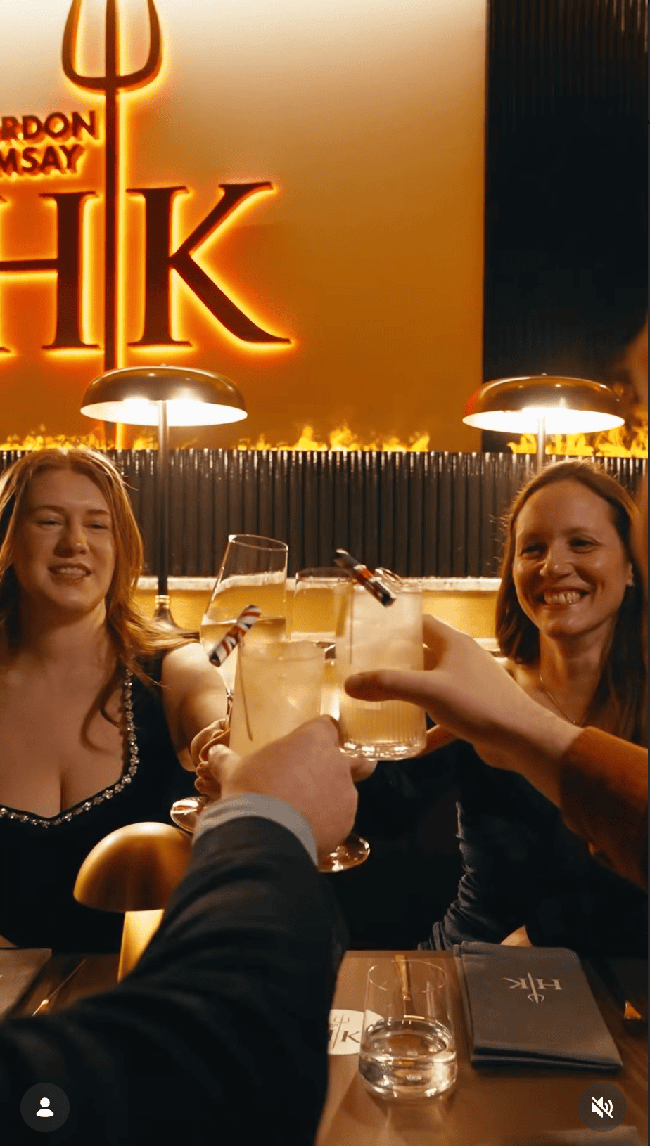 Four people clink glasses at a restaurant table under a glowing “Gordon Ramsay HK” sign, smiling.