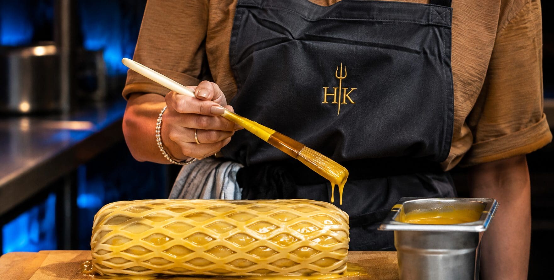 A person brushes egg wash on a lattice pastry in a kitchen, wearing an apron with the HK logo.