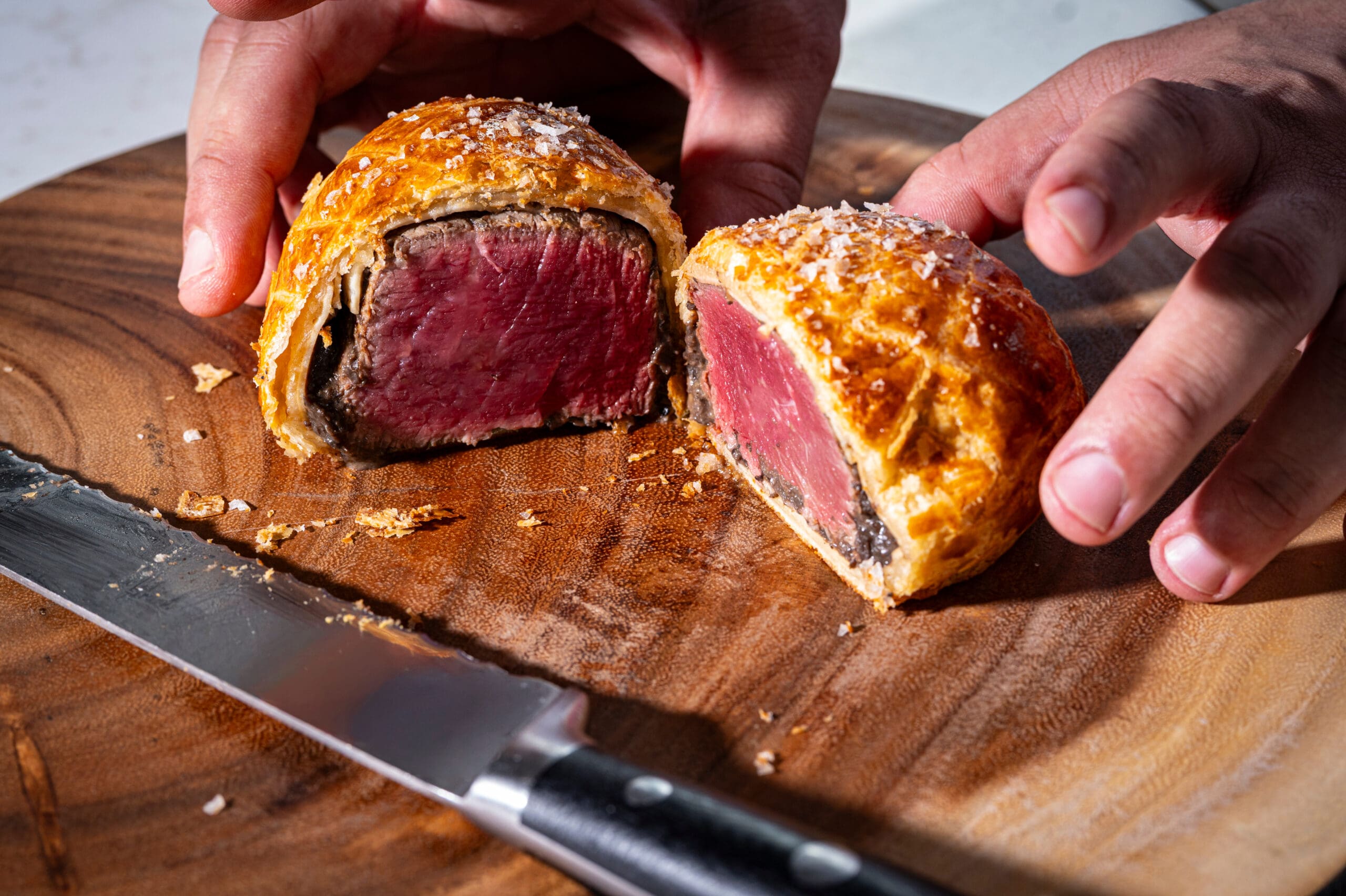 Hands holding a sliced beef Wellington on a wooden board, with a knife beside it.
