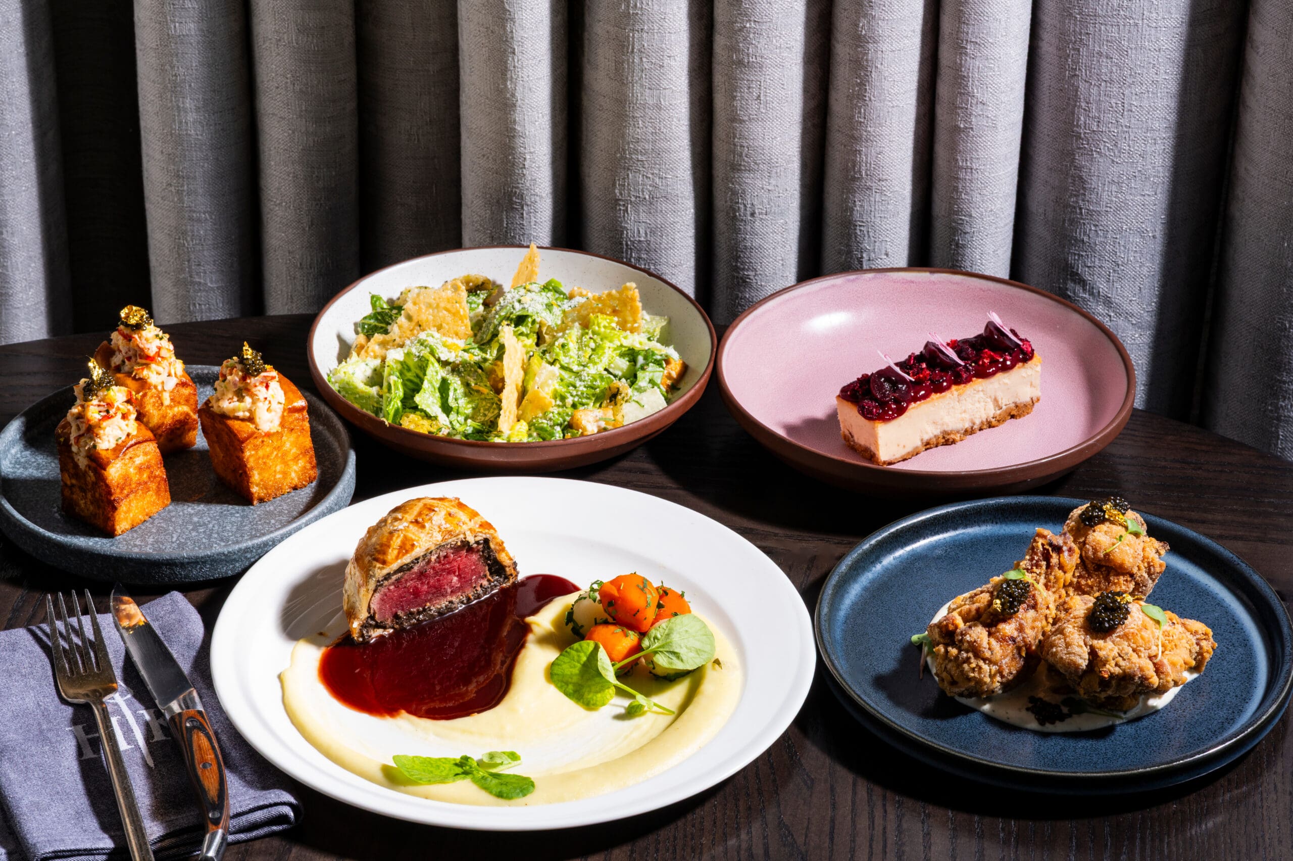 Five plated gourmet dishes, including salad, steak, fried chicken, cake, and cornbread, on a dark table.