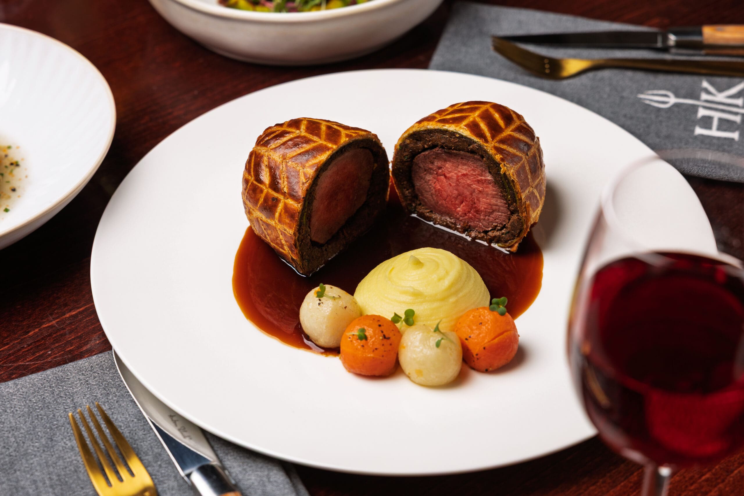 Beef Wellington with mashed potato, glazed vegetables, and sauce, served on a white plate with red wine.