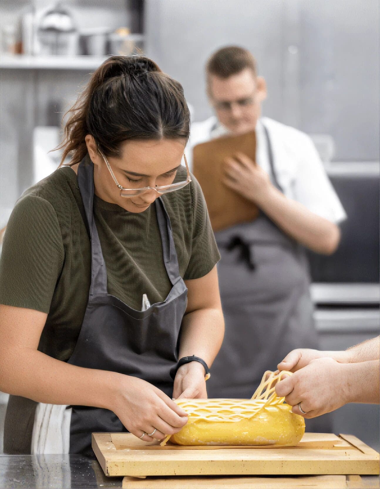 Woman in apron decorates pastry with lattice pastry; another person observes in kitchen background.