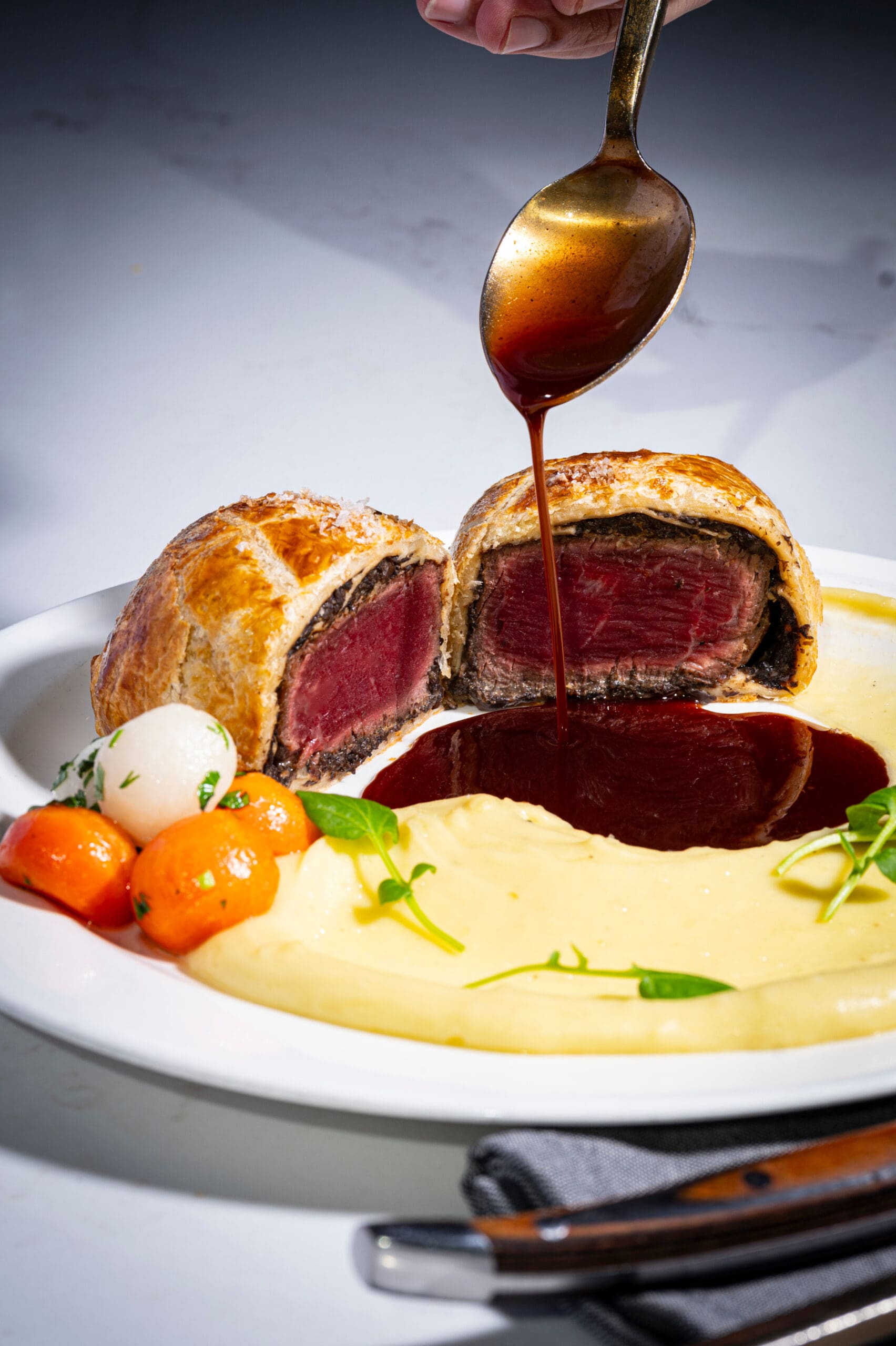 Sliced beef Wellington with mashed potatoes, glazed vegetables, and sauce being poured on a white plate.