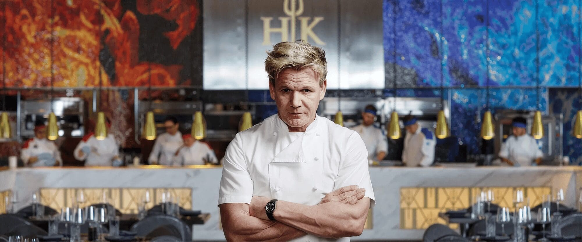 Chef in white stands with arms folded in a restaurant kitchen, staff working in the background.