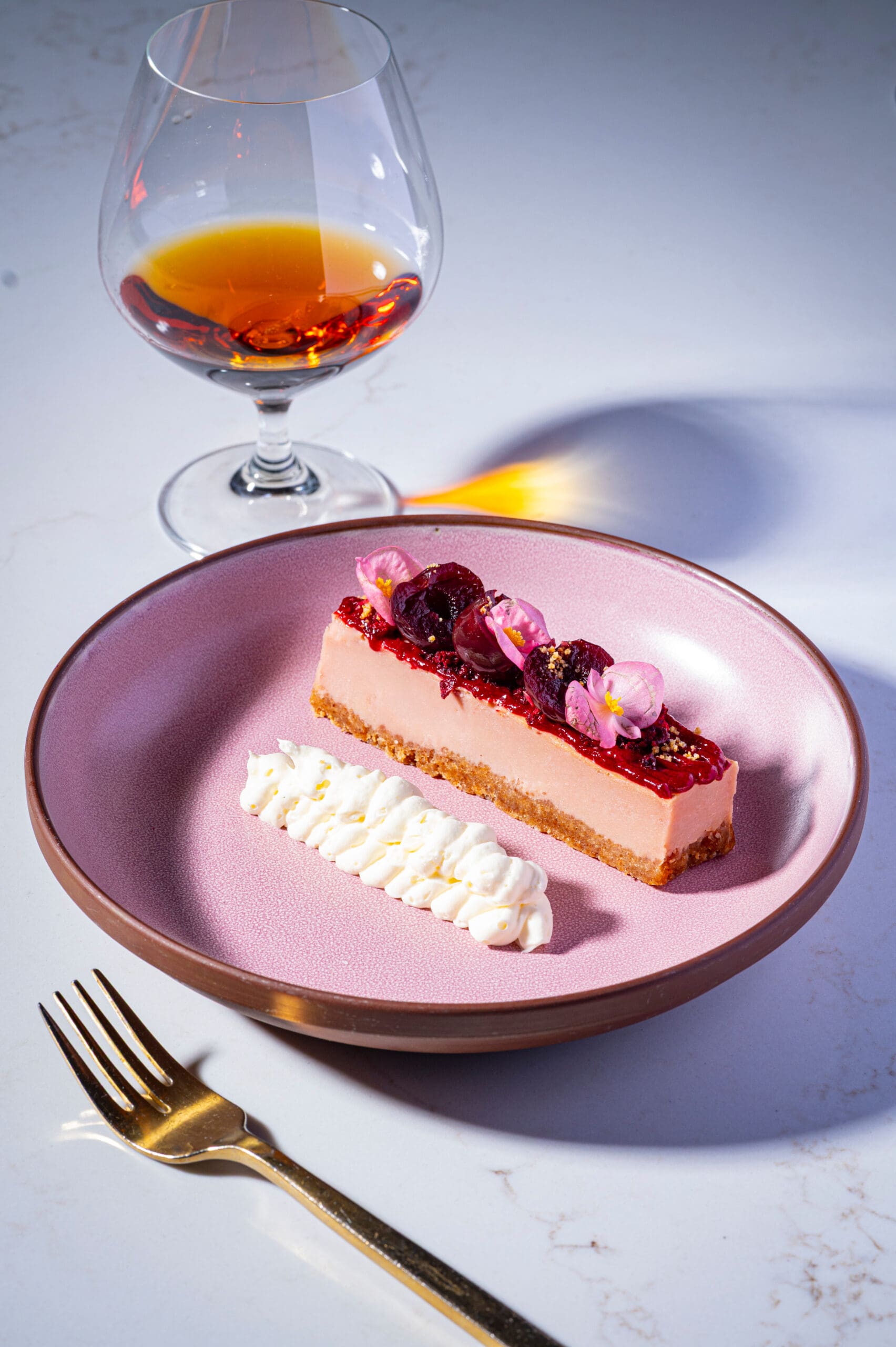 Pink dessert with whipped cream, edible flowers, and a drink in a glass on a white table with a gold fork.