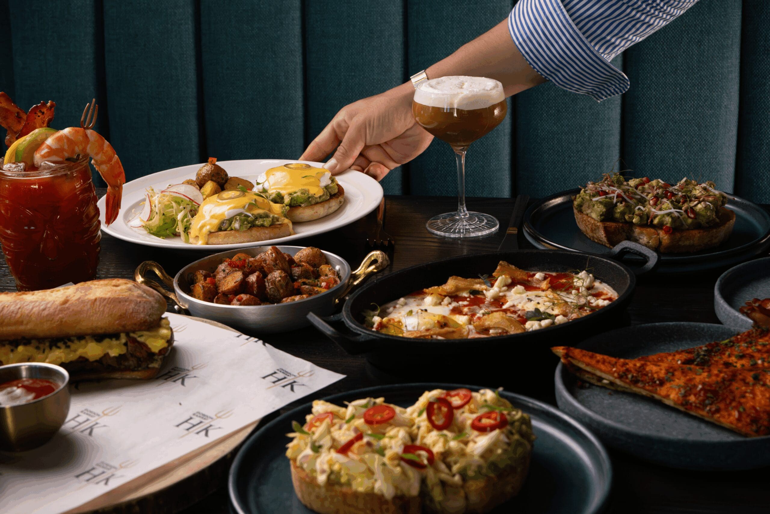 A hand reaches for a plate of eggs Benedict among various brunch dishes and drinks on a table.