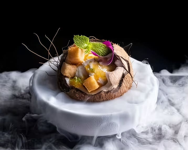 A gourmet dessert in a coconut shell on a smoky, misty plate, garnished with mint and edible flowers.