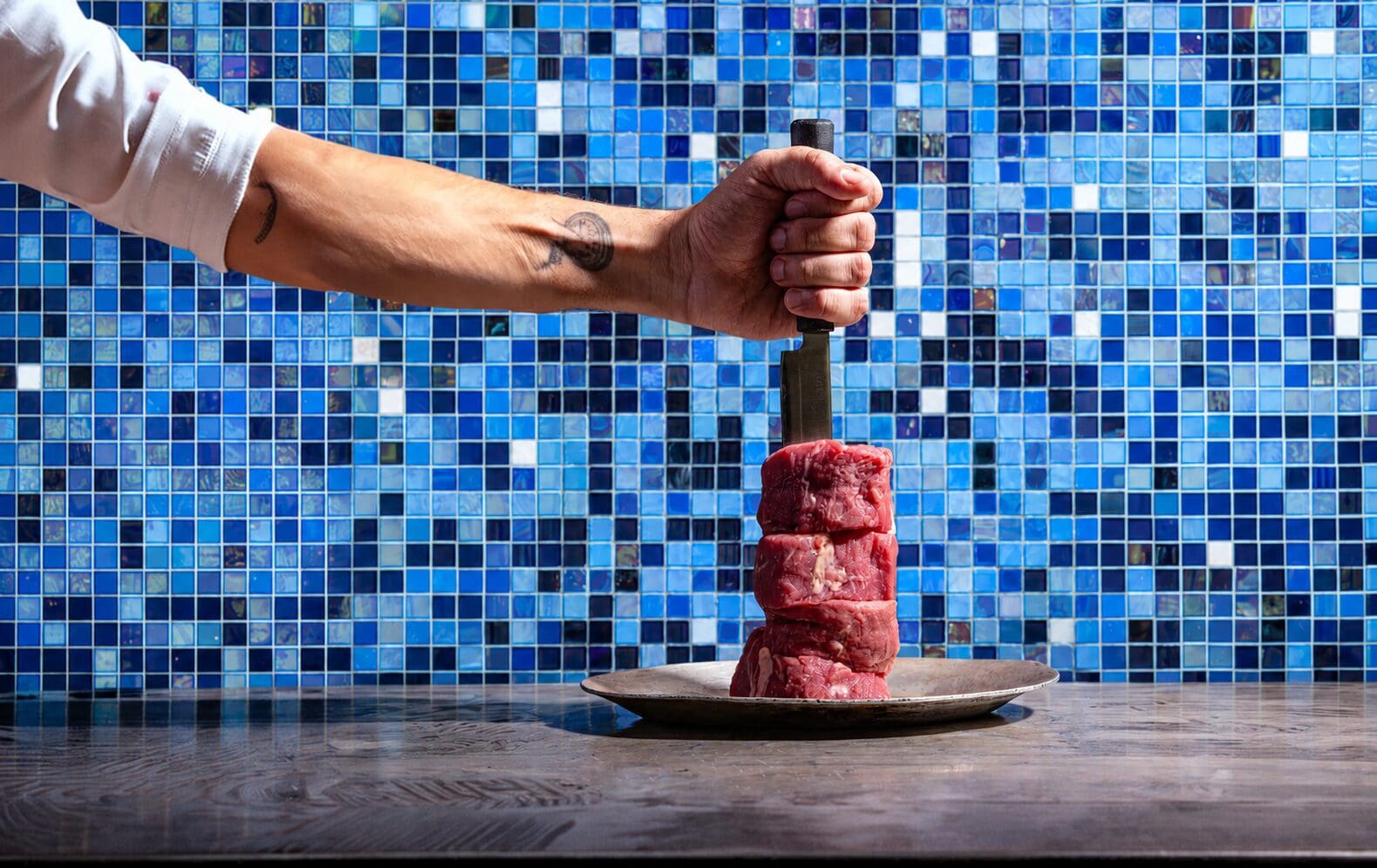 A hand grips a knife stabbed into stacked raw meat on a plate, with a blue tiled wall in the background.